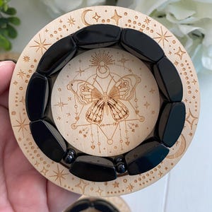 May include: A stretch bracelet composed of smooth, rectangular black beads resting on a circular wooden disc. The wood features a laser-engraved design of a butterfly centered within geometric lines, surrounded by stars, moons, and dots.