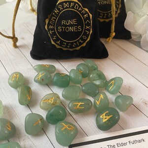 May include: A collection of green rune stones with gold lettering, scattered near two black velvet drawstring bags. The bags are labeled "RUNE STONES" in gold lettering. A reference card with rune symbols is also present.