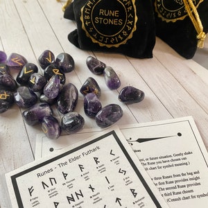 May include: Amethyst rune stones with gold symbols are scattered near black velvet bags labeled "Rune Stones." A chart of Elder Futhark runes is also present. The stones are a deep purple color.