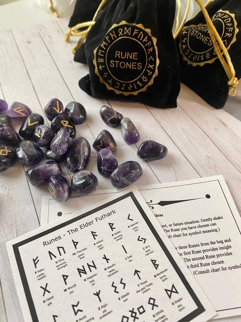 May include: Amethyst rune stones with gold symbols are scattered near black velvet bags labeled "Rune Stones." A chart of Elder Futhark runes is also present. The stones are a deep purple color.