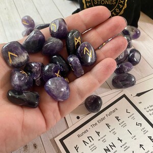 May include: Amethyst rune stones with gold symbols are held in a hand. The stones are next to a black velvet bag with "RUNE STONES" in gold lettering. A chart of the Elder Futhark runes is also visible.