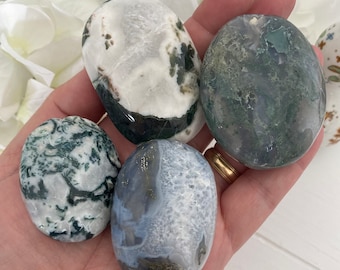May include: Four polished stones in shades of white, green, and blue are held in a hand. The stones have unique patterns and textures, with some featuring moss-like inclusions. The background includes white flowers.