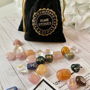 May include: A black velvet drawstring bag with gold embroidered text reading "RUNE STONES" sits among a collection of polished rune stones. The stones are various colors, including clear, pink, and yellow, with gold rune symbols. A card with Elder Futhark runes is also present.