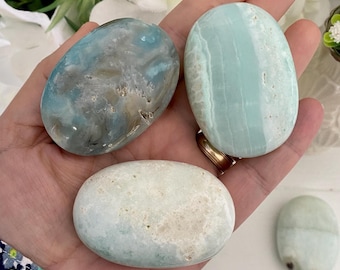 May include: A hand holds several smooth, oval-shaped stones. The stones vary in color, including shades of blue, green, and white, with natural patterns and textures. The stones are likely used for decorative or therapeutic purposes.