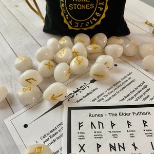 May include: A collection of white rune stones with gold symbols, scattered on a light wood surface. A black velvet bag with gold lettering that reads "Rune Stones" is in the background, along with a chart of runes.