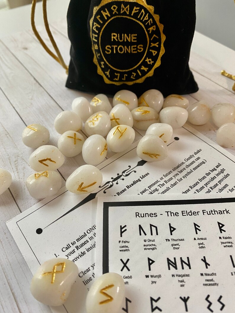 May include: A collection of white rune stones with gold symbols, scattered on a light wood surface. A black velvet bag with gold lettering that reads "Rune Stones" is in the background, along with a chart of runes.
