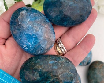 May include: A collection of polished, oval-shaped blue apatite stones. The stones have a mottled appearance with shades of teal, navy, and brown. The stones are smooth and appear to be natural gemstones. The background includes white flowers and a hand.
