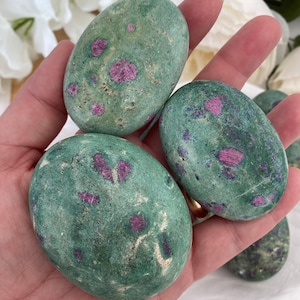 May include: A hand holds several oval-shaped stones. The stones are a mottled green color with small, irregular patches of deep pink. The stones have a smooth, polished appearance. The background is blurred, with white flowers visible.