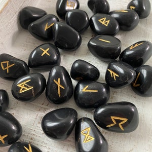 May include: A collection of polished black stones, each engraved with a gold rune symbol. The stones are smooth and rounded, resting on a light-colored wooden surface. The runes are in a gold color.