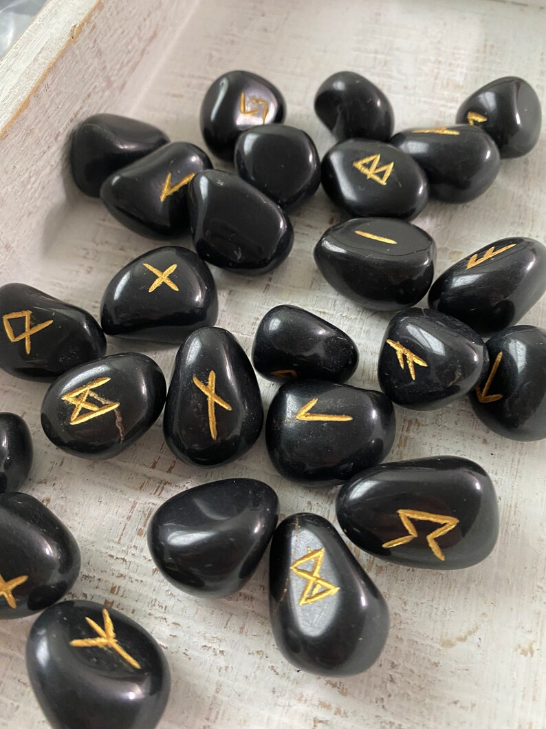 May include: A collection of polished black stones, each engraved with a gold rune symbol. The stones are smooth and rounded, resting on a light-colored wooden surface. The runes are in a gold color.