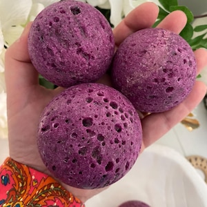 May include: Three round, porous, purple stones are held in a hand. The stones have a textured surface with small holes. The background includes white flowers and green leaves. The stones are a deep shade of purple.