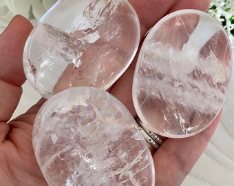 May include: Three clear quartz palm stones with a smooth, polished surface. The stones are oval-shaped and held in a hand, showcasing their translucent quality and subtle internal patterns. The background features white flowers.