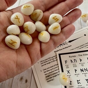 May include: A collection of white, oval-shaped rune stones with gold-colored symbols. The stones are held in a hand, with a printed guide and instructions visible in the background. The runes are for divination and spiritual guidance.