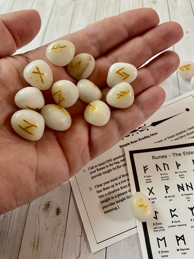 May include: A collection of white, oval-shaped rune stones with gold-colored symbols. The stones are held in a hand, with a printed guide and instructions visible in the background. The runes are for divination and spiritual guidance.