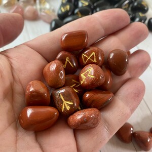 May include: A handful of polished, reddish-brown rune stones with gold-colored symbols. Other stones in shades of pink, brown, and black are visible in the background. The stones are smooth and appear to be made of natural materials.