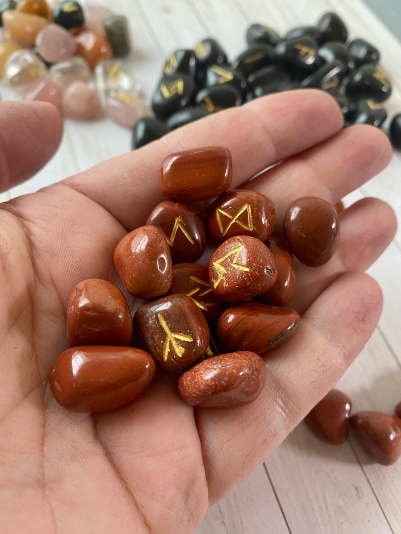 May include: A handful of polished, reddish-brown rune stones with gold-colored symbols. Other stones in shades of pink, brown, and black are visible in the background. The stones are smooth and appear to be made of natural materials.
