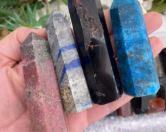Peut inclure: Quatre tours de cristal polies tenues dans une paume. La plus à gauche est rose et grise tachetée, la deuxième est grise avec des bandes bleues, la troisième est une pierre noire haute et lisse avec une fissure, et la dernière est bleu vif.