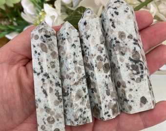 May include: Four polished stone towers with a speckled pattern of gray, brown, and black on a white and light green background. The towers are held in a hand, with a blurred floral arrangement in the background.