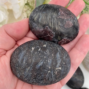 May include: Two oval-shaped, polished stones with a dark, charcoal-gray base color. The stones have a textured, fibrous pattern with streaks of silver and burgundy. The stones are held in an open hand, with a blurred floral background.