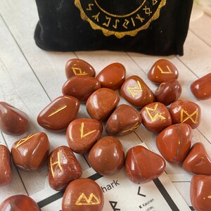 May include: A collection of polished, reddish-brown rune stones with gold-colored symbols. A black velvet bag with gold lettering that reads "RUNE STONES" is in the background. Cards with rune symbols are also visible.