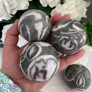 May include: Four decorative stone spheres with a marbled pattern of gray and white swirls. One sphere rests in a white, shell-shaped dish. The spheres are held in a hand, with white flowers in the background.