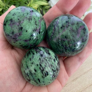May include: Three polished, round stones with a green base color, speckled with black and purple inclusions. The stones are smooth and spherical, held in an open hand. The background features greenery and white flowers.