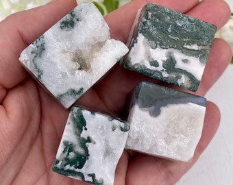 May include: Four moss agate cubes, each displaying a unique pattern of white and green. The cubes are held in a hand, showcasing their natural textures and colors. The moss agate has a crystalline structure.