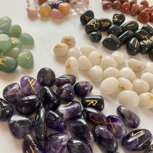 May include: Assorted tumbled stones in various colors, including clear, pink, green, black, white, and purple. Each stone features a gold-colored rune symbol. The stones are arranged in small piles, showcasing their different shapes and textures.