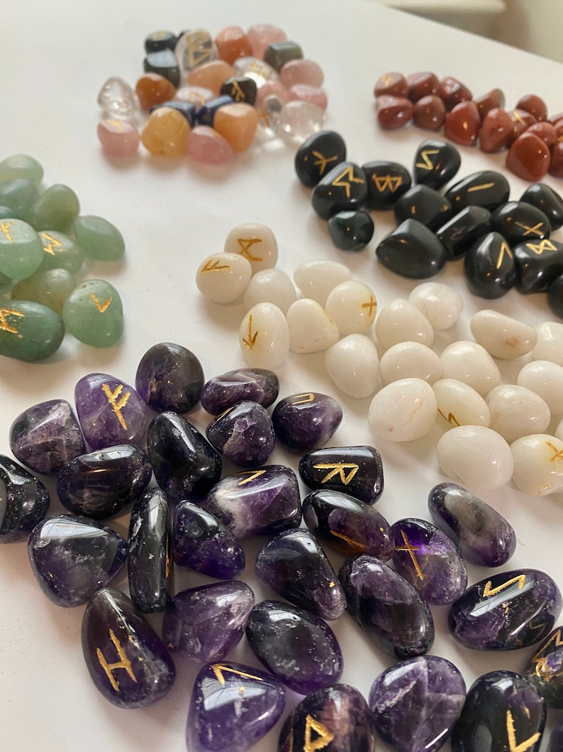 May include: Assorted tumbled stones in various colors, including clear, pink, green, black, white, and purple. Each stone features a gold-colored rune symbol. The stones are arranged in small piles, showcasing their different shapes and textures.