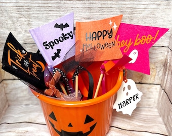 May include: An orange plastic jack-o'-lantern bucket filled with Halloween-themed decorations. The bucket contains several colorful pennant flags with phrases like "Spooky," "Happy Halloween," and "hey boo!" A white ghost-shaped tag with the name "Harper" hangs from the bucket.