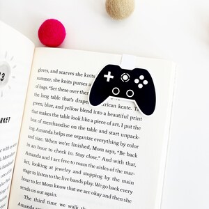 May include: A black and white bookmark shaped like a video game controller. The bookmark is on a page of a book with text about a table draped in green, blue, and yellow fabric. The text reads "Set these over there...the long table that's draped in green, blue, and yellow blend into a beautiful print that makes the table look like a piece of art."