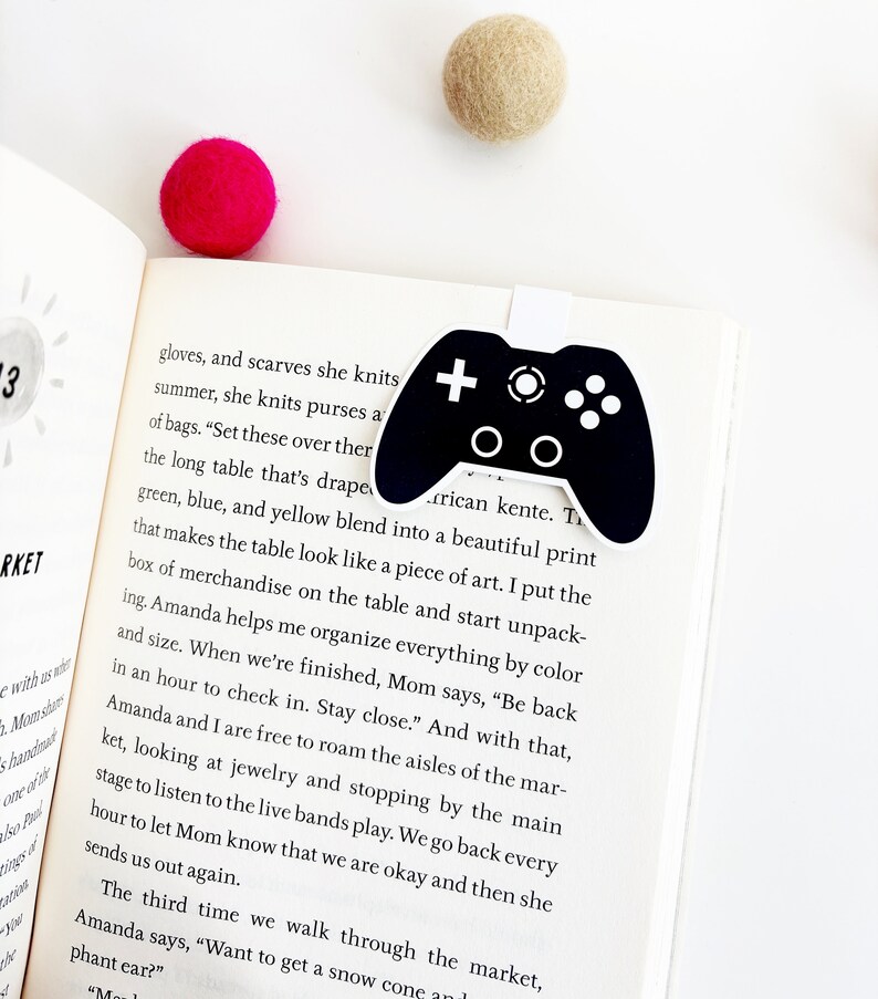 May include: A black and white bookmark shaped like a video game controller. The bookmark is on a page of a book with text about a table draped in green, blue, and yellow fabric. The text reads "Set these over there...the long table that's draped in green, blue, and yellow blend into a beautiful print that makes the table look like a piece of art."