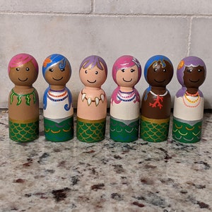 May include: Six hand-painted wooden mermaid peg dolls. Each doll has a different skin tone, hair color, and necklace. The mermaid tails are green with gold accents. The dolls are standing on a speckled countertop.