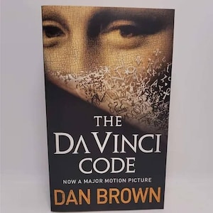 May include: A paperback book cover with a close-up of the Mona Lisa's eye. The title is "The Da Vinci Code" with the text "Now a Major Motion Picture" and the author's name "Dan Brown" in orange.
