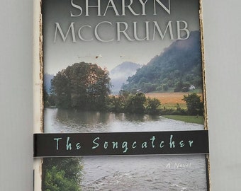 May include: Book cover featuring a landscape scene with a river, trees, and mountains. The title "The Songcatcher" is written in a cursive font on a black band across the bottom of the image. The author's name, "Sharyn McCrumb", is printed in large, bold letters above the image. The text "New York Times Bestselling Author" is printed in smaller letters above the author's name. The text "A Novel" is printed in smaller letters below the title.