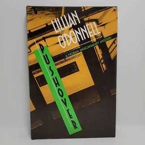 May include: A paperback book titled "Pushhover" by Lillian O'Donnell. The book cover features a yellow and black building with a green stripe that reads "Pushhover" in black letters. The book is a Norah Mulcahaney mystery.