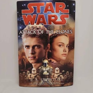 May include: A book cover for the Star Wars Episode II: Attack of the Clones novel. The cover features a red and white title with a background image of Anakin Skywalker and Padmé Amidala. The book is written by R.A. Salvatore.