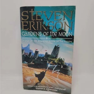 K&ouml;nnte beinhalten: Ein Taschenbuchcover mit blauem und gr&uuml;nem Hintergrund. Der Titel lautet "Gardens of the Moon" von Steven Erikson. Der Untertitel lautet "A Tale of the Malazan Book of the Fallen". Das Cover zeigt eine Fantasy-Illustration eines Zauberers, der an einem Strand steht, mit einer Stadt im Hintergrund.