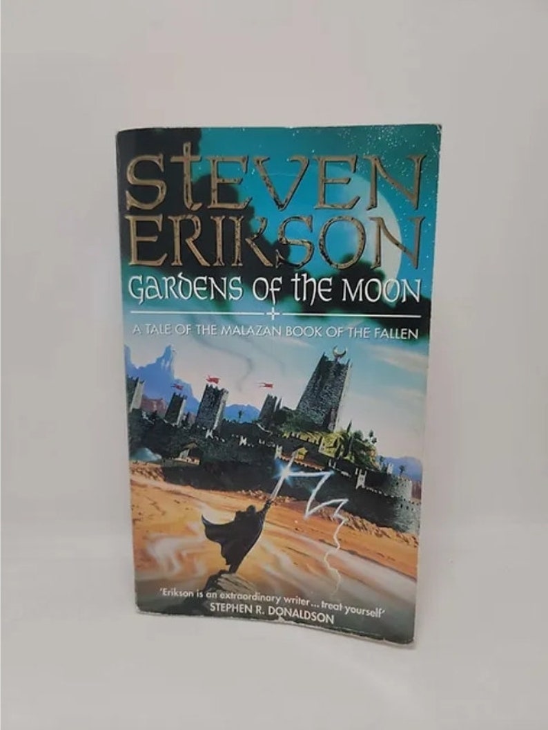 K&ouml;nnte beinhalten: Ein Taschenbuchcover mit blauem und gr&uuml;nem Hintergrund. Der Titel lautet "Gardens of the Moon" von Steven Erikson. Der Untertitel lautet "A Tale of the Malazan Book of the Fallen". Das Cover zeigt eine Fantasy-Illustration eines Zauberers, der an einem Strand steht, mit einer Stadt im Hintergrund.