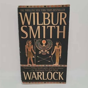 May include: A black and gold paperback book with the title "WARLOCK" by Wilbur Smith. The cover features an illustration of two figures in ancient Egyptian clothing, a bird, and two arrows.