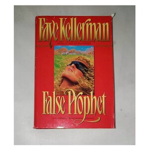 May include: A red paperback book with the title "False Prophet" by Faye Kellerman, author of "Day of Atonement". The book cover features a woman with her eyes covered by a blindfold, lying on her back with her head tilted back.