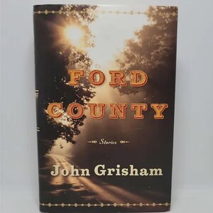 May include: A book cover with a photo of a country road leading into a forest with a bright sun shining through the trees. The title "FORD COUNTY" is in large, orange letters with a gold outline. The author's name "John Grisham" is below the title in smaller, black letters. The book cover also includes the words "Stories" in smaller, black letters.