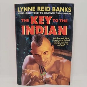 May include: Book cover for "The Key to the Indian" by Lynne Reid Banks, a novel about a young boy who travels back in time to help a Native American. The cover features a Native American man with a mohawk and a serious expression, holding his hands out in front of him. The book title is in large, bold letters.