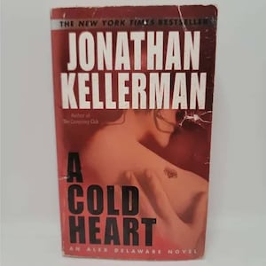 May include: A paperback book cover with a red background. The title is "A Cold Heart" and the author is "Jonathan Kellerman". The book is part of the "Alex Delaware" series.