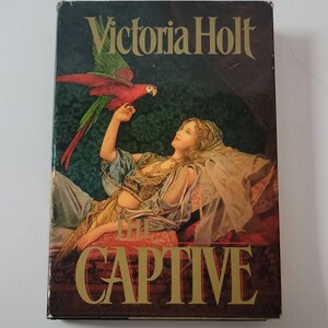 May include: A book cover with a dark background featuring the title "The Captive" in gold lettering. The author, Victoria Holt, is printed in white lettering above the title. The cover art depicts a woman in a blue and gold dress lying on a couch with a red and green parrot perched on her hand.