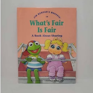 May include: A pink book with the title "What's Fair Is Fair" and a subtitle "A Book About Sharing". The book features illustrations of Kermit the Frog and Miss Piggy riding a Ferris wheel.