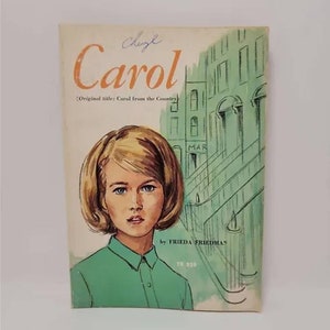 May include: A vintage paperback book cover with a green background featuring a young woman with blonde hair and a green shirt. The title of the book is "Carol" with the original title "Carol from the Country" in parentheses. The author is "Frieda Friedman" and the book number is "TX 859".