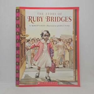 May include: A Scholastic book cover featuring an illustration of a young girl, Ruby Bridges, walking into a school building. The cover is red with the title "The Story of Ruby Bridges" in white text. The book is part of the Scholastic Book Fairs series.