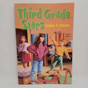 May include: A book cover with the title "Third Grade Stars" by Candice F. Ransom. The cover features three children in a workshop setting. A girl in a pink jacket stands with her hands on her hips, a girl in a red skirt and white shirt looks surprised, and a boy in a yellow shirt balances on a wooden plank.