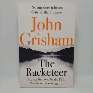 May include: A book cover with a black and white image of a lake and trees. The title is "The Racketeer" by John Grisham. The text on the cover reads "He was betrayed by the FBI. Now he wants revenge..."